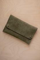 An olive green coloured suede clutch with small gold coloured metal studs, displayed diagonally on a light wood grain surface.