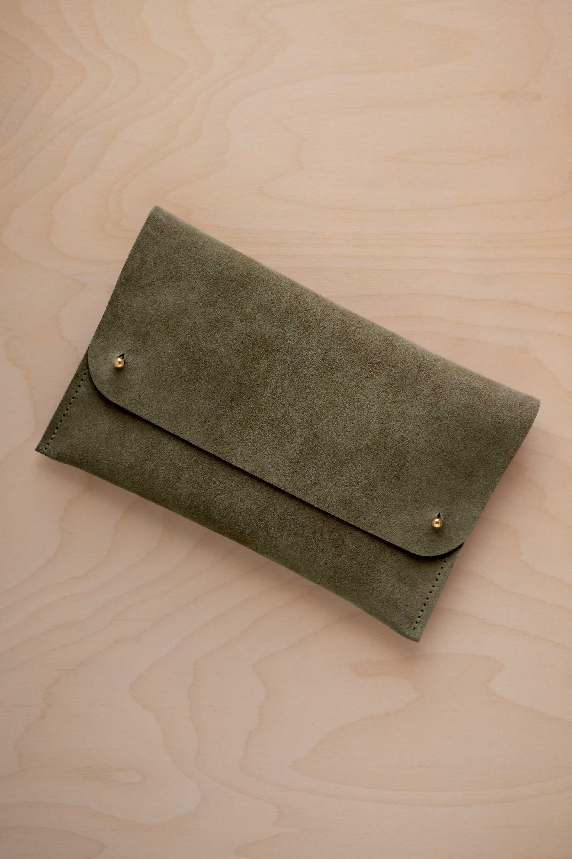 An olive green coloured suede clutch with small gold coloured metal studs, displayed diagonally on a light wood grain surface.