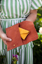 A person in a green and white striped dress holds an open terracotta brown suede pouch or clutch bag, showing a small yellow envelope-style purse inside. The background is a softly blurred green outdoor setting with a purple flower in the foreground.