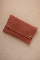A terracotta coloured suede clutch with small gold coloured metal studs, displayed diagonally on a light wood grain surface.