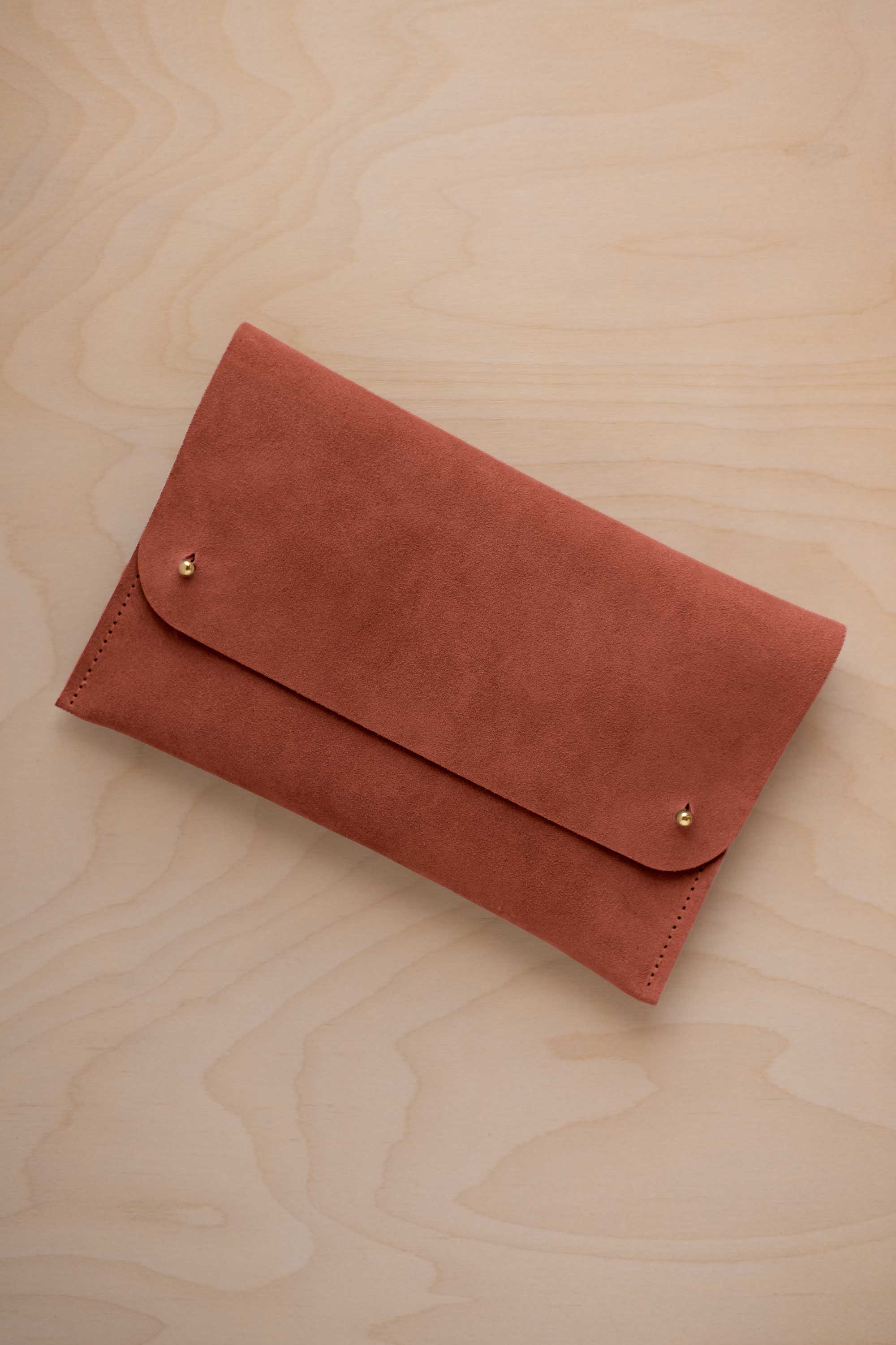 A terracotta coloured suede clutch with small gold coloured metal studs, displayed diagonally on a light wood grain surface.