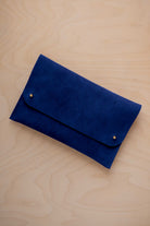 An ultramarine blue coloured suede clutch with small gold coloured metal studs, displayed diagonally on a light wood grain surface.