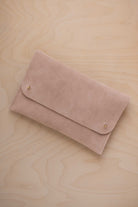 A warm beige suede clutch with small gold coloured metal studs, displayed diagonally on a light wood grain surface.