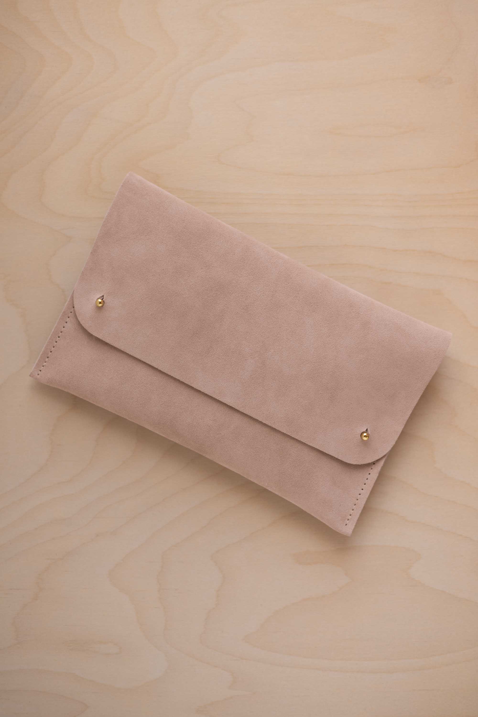 A warm beige suede clutch with small gold coloured metal studs, displayed diagonally on a light wood grain surface.