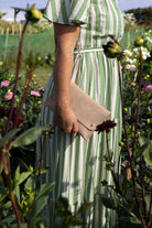 A person standing in a flower garden wearing a green and white vertically striped midi dress and holding a beige suede clutch bag.