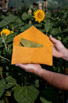 A mustard yellow-orange suede envelope-style pouch with a flap closure is held open by two hands in a sunflower field. A green suede purse is tucked inside the pouch.