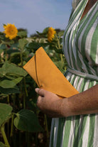 A person in a green and white striped dress holding a bright yellow-orange envelope-style clutch in a sunflower field.