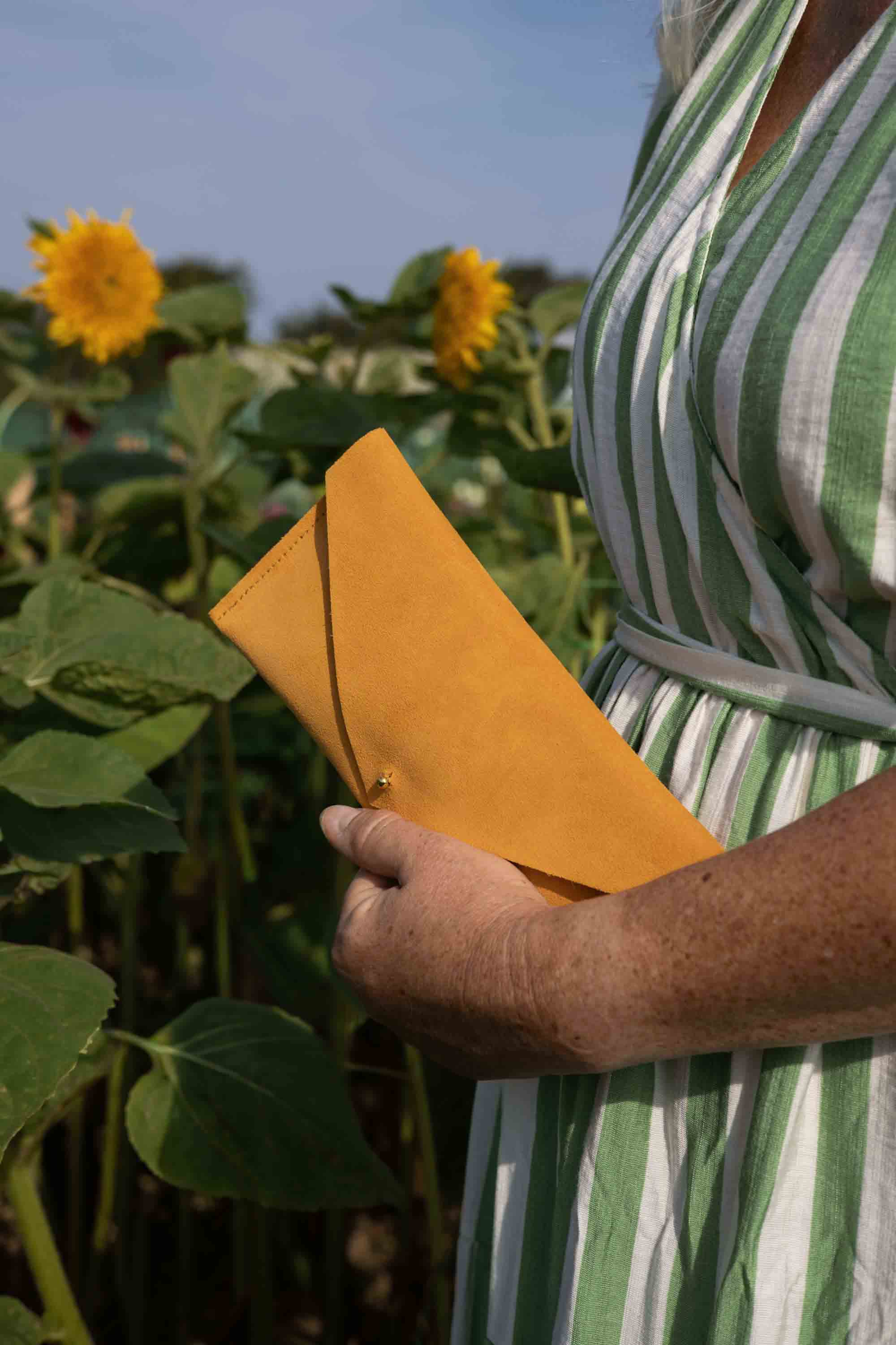 A person in a green and white striped dress holding a bright yellow-orange envelope-style clutch in a sunflower field.