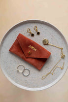 A top-down view of a terracotta coloured suede jewellery pouch with gold foiled initials with a gold coloured fastening, displayed on a round speckled ceramic plate alongside gold and silver jewellery pieces on a warm beige background.