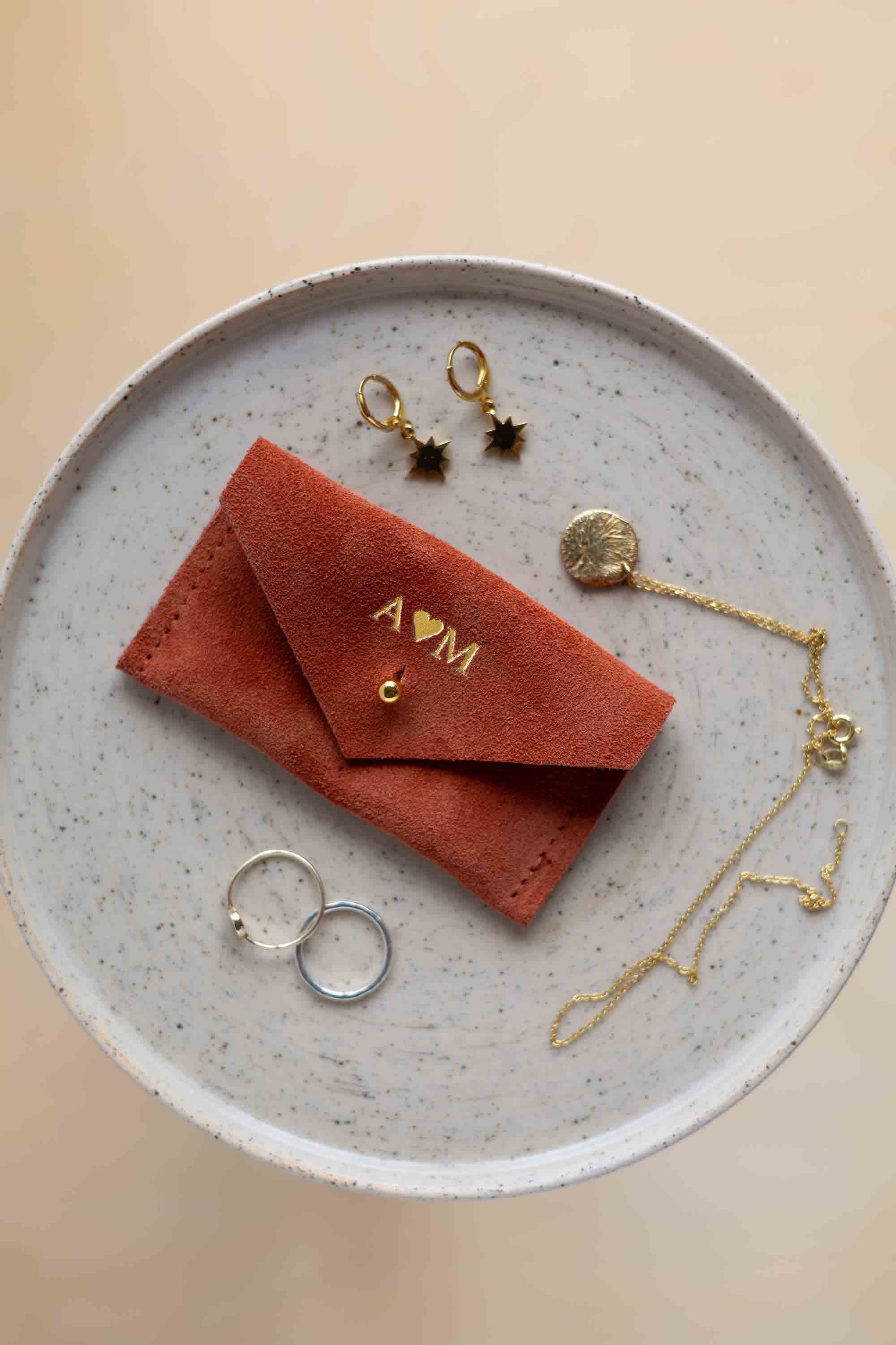 A top-down view of a terracotta coloured suede jewellery pouch with gold foiled initials with a gold coloured fastening, displayed on a round speckled ceramic plate alongside gold and silver jewellery pieces on a warm beige background.