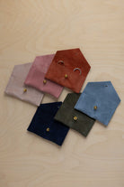 Six small suede pouches in assorted colours arranged together on a light wood background. The rust coloured suede pouch has couples wedding rings nestled inside.