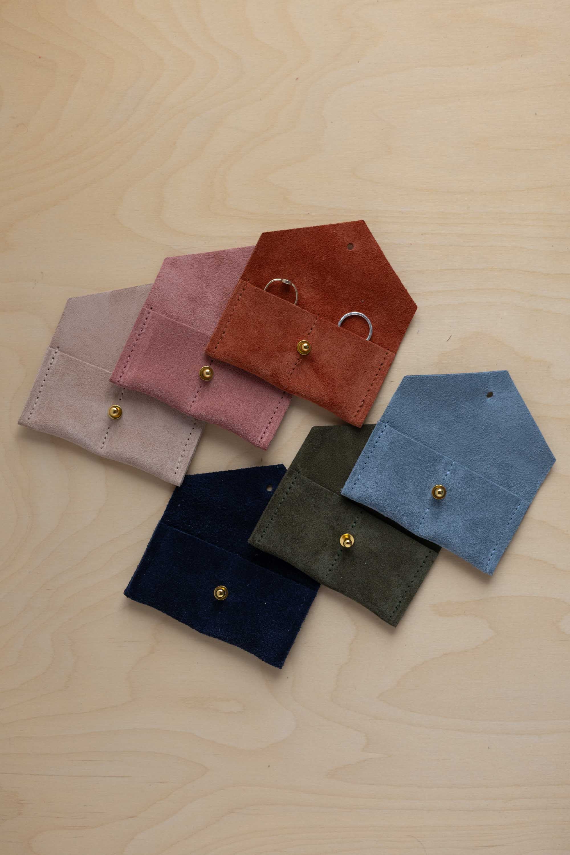 Six small suede pouches in assorted colours arranged together on a light wood background. The rust coloured suede pouch has couples wedding rings nestled inside.