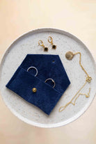 A top-down view of an ultramarine blue coloured suede jewellery pouch with a gold coloured fastening, displayed on a round speckled ceramic plate alongside gold and silver jewellery pieces on a warm beige background.