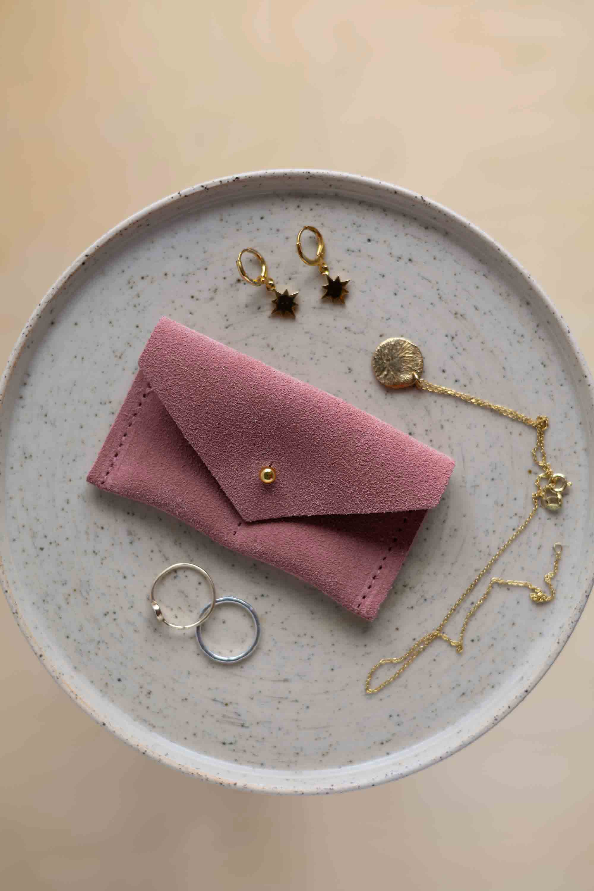A top-down view of a dusky pink suede jewellery pouch with a gold coloured fastening, displayed on a round speckled plate alongside gold and silver jewellery pieces on a warm beige background.