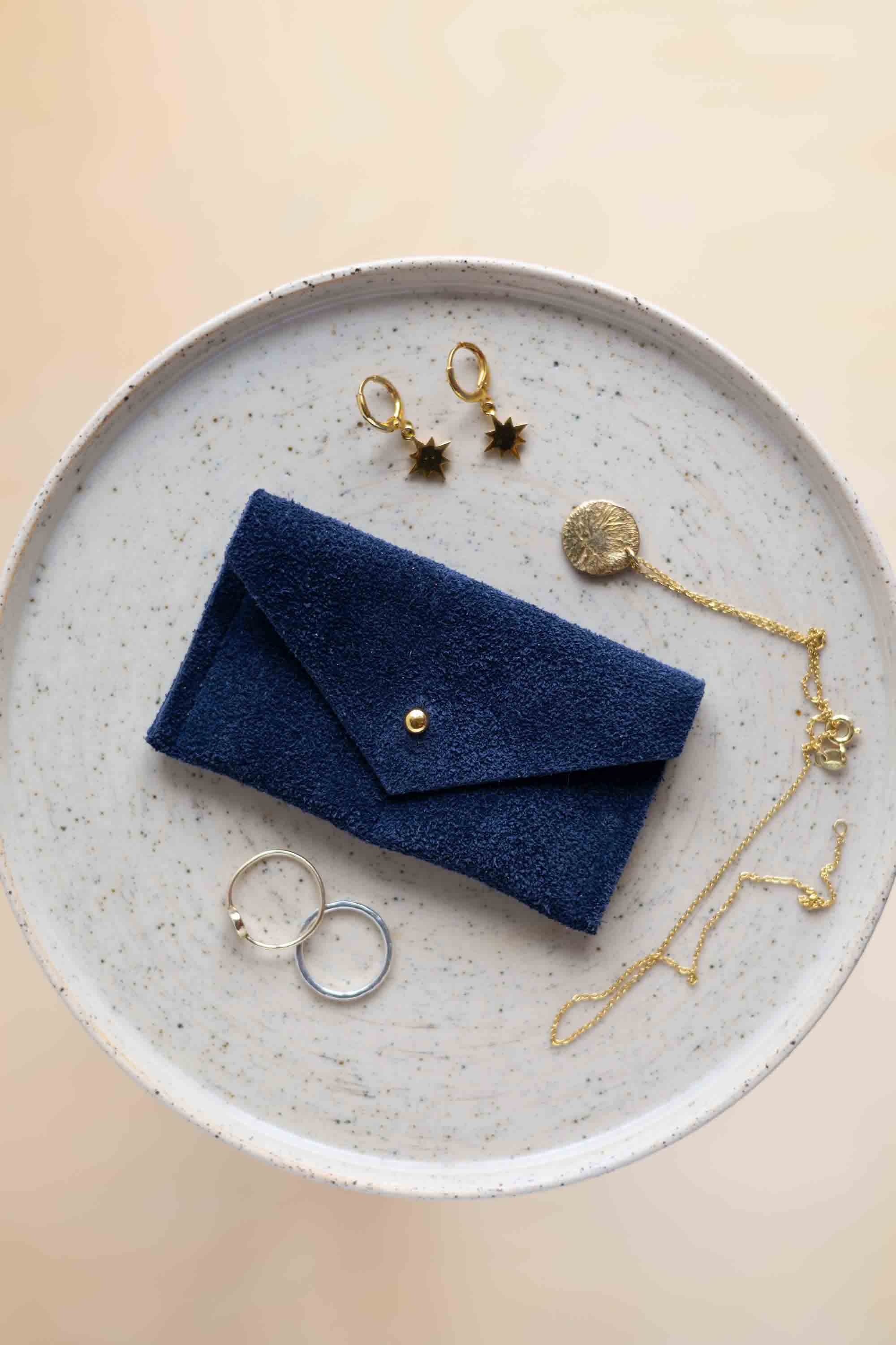 A top-down view of a midnight navy suede jewellery pouch with a gold coloured fastening, displayed on a round speckled ceramic plate alongside gold and silver jewellery pieces on a warm beige background.