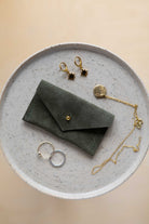 A top-down view of a soft olive green suede jewellery pouch with a gold coloured fastening, displayed on a round speckled ceramic plate alongside old and silver jewellery pieces on a warm beige background.