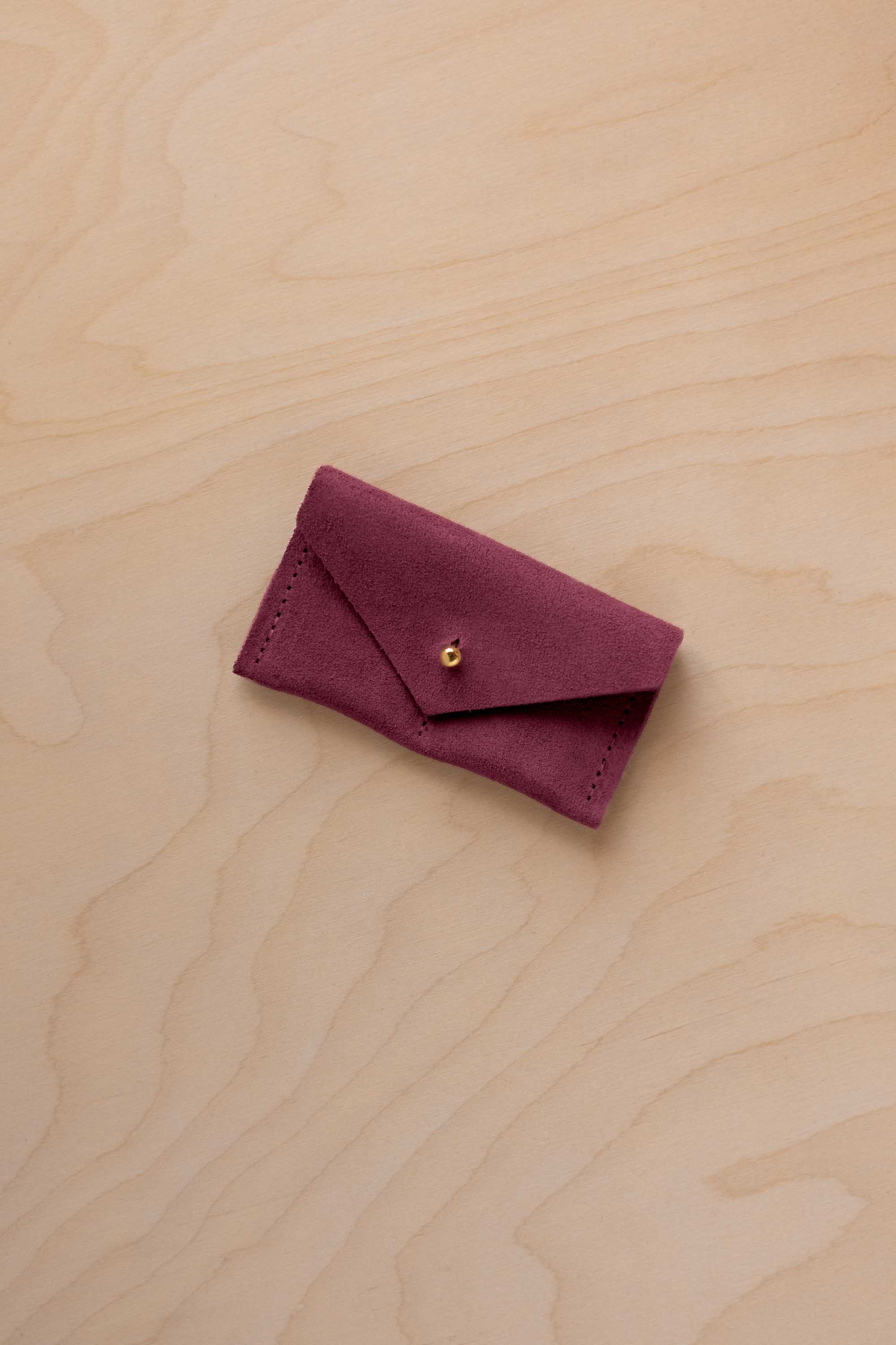 A small burgundy coloured suede jewellery pouch with a gold coloured fastening, resting on a light wood grain surface.