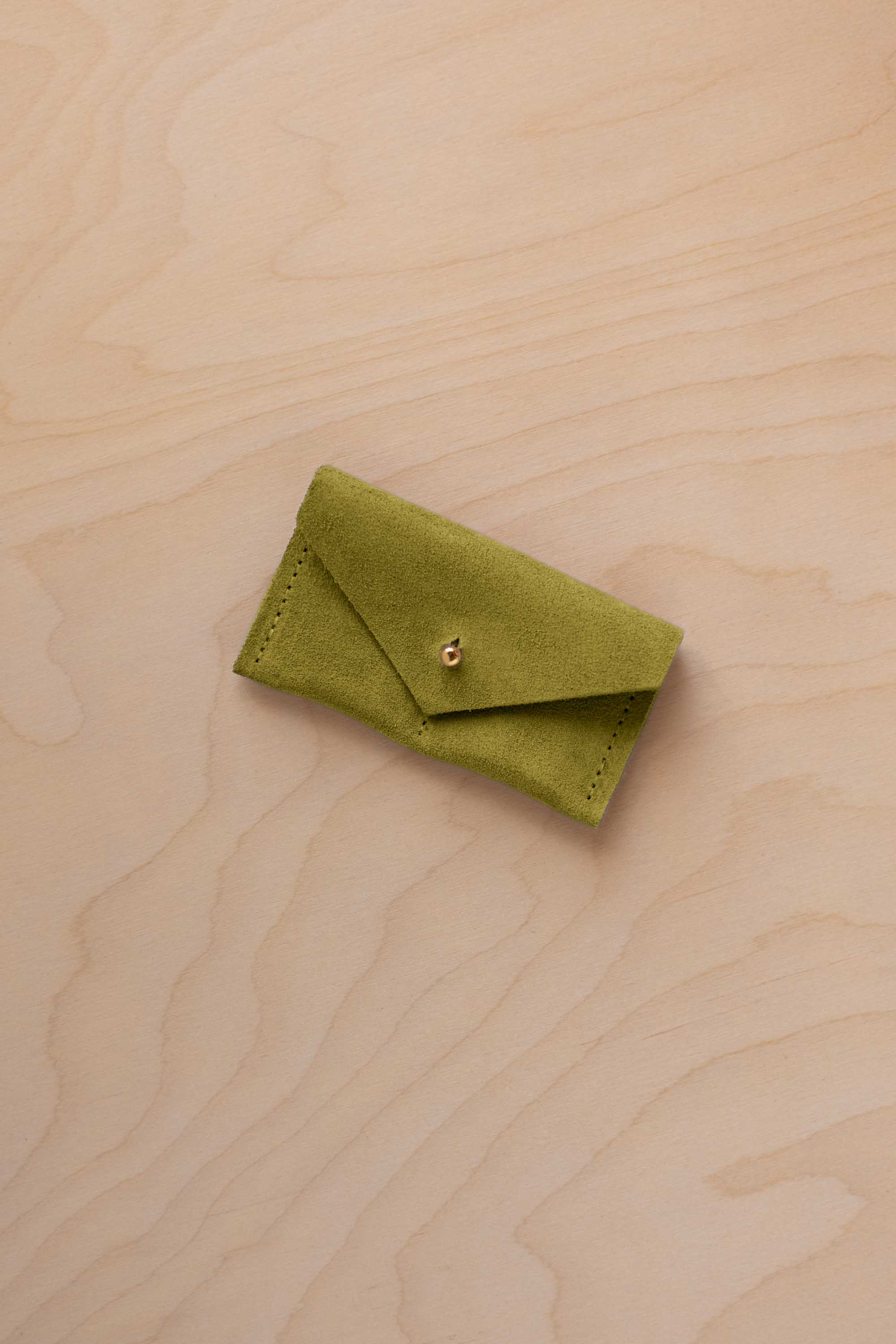 A small chartreuse green coloured suede jewellery pouch with a gold button, resting on a light wood grain surface.
