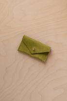A small chartreuse green coloured suede jewellery pouch with a gold button, resting on a light wood grain surface.