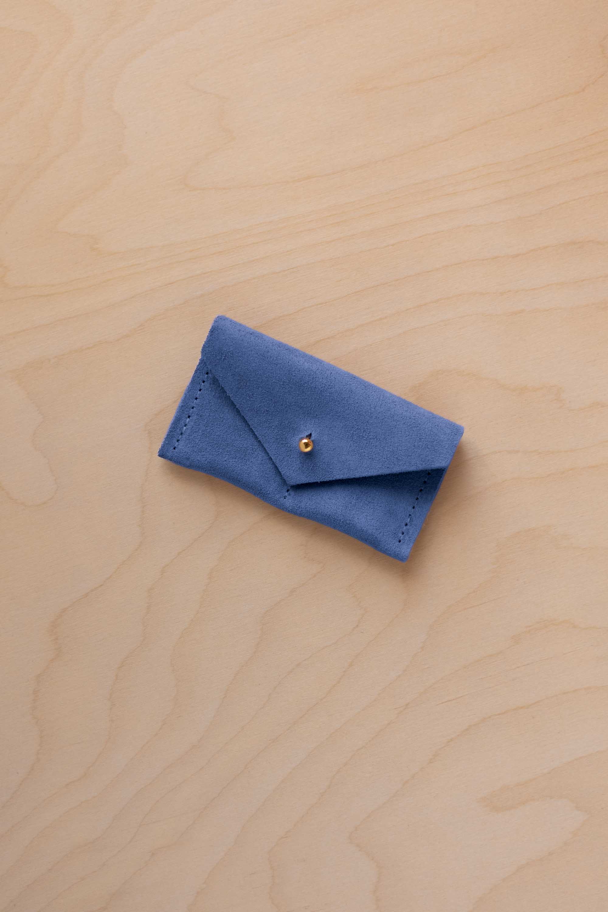 A small cobalt blue coloured suede jewellery pouch with a gold coloured fastening, resting on a light wood grain surface.
