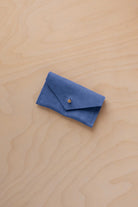 A small cobalt blue coloured suede jewellery pouch with a gold coloured fastening, resting on a light wood grain surface.