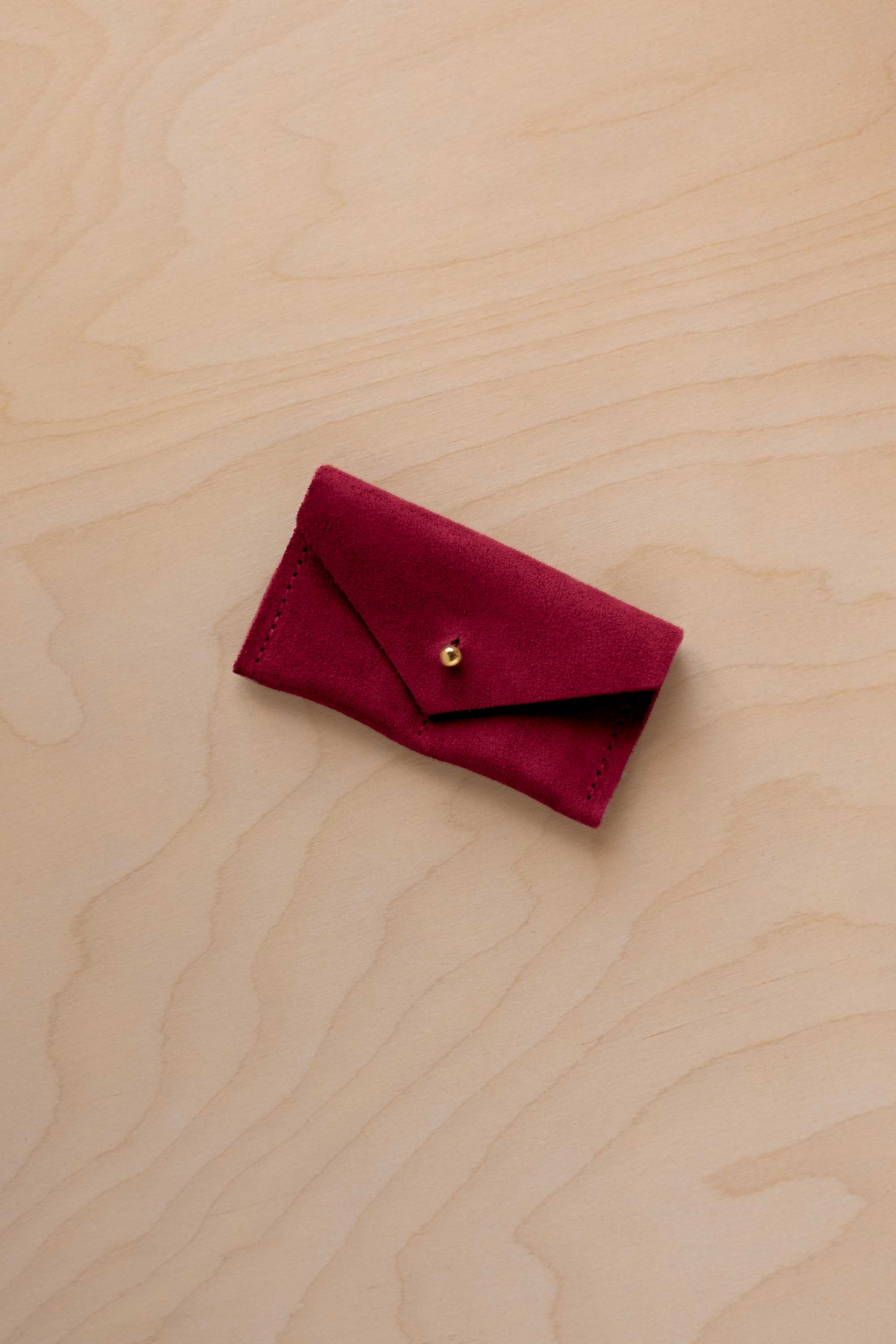 A small crimson red coloured suede jewellery pouch with a gold coloured fastening, resting on a light wood grain surface.
