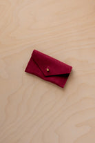 A small crimson red coloured suede jewellery pouch with a gold coloured fastening, resting on a light wood grain surface.