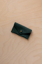 A small deep forest coloured suede jewellery pouch with a gold button, resting on a light wood grain surface.