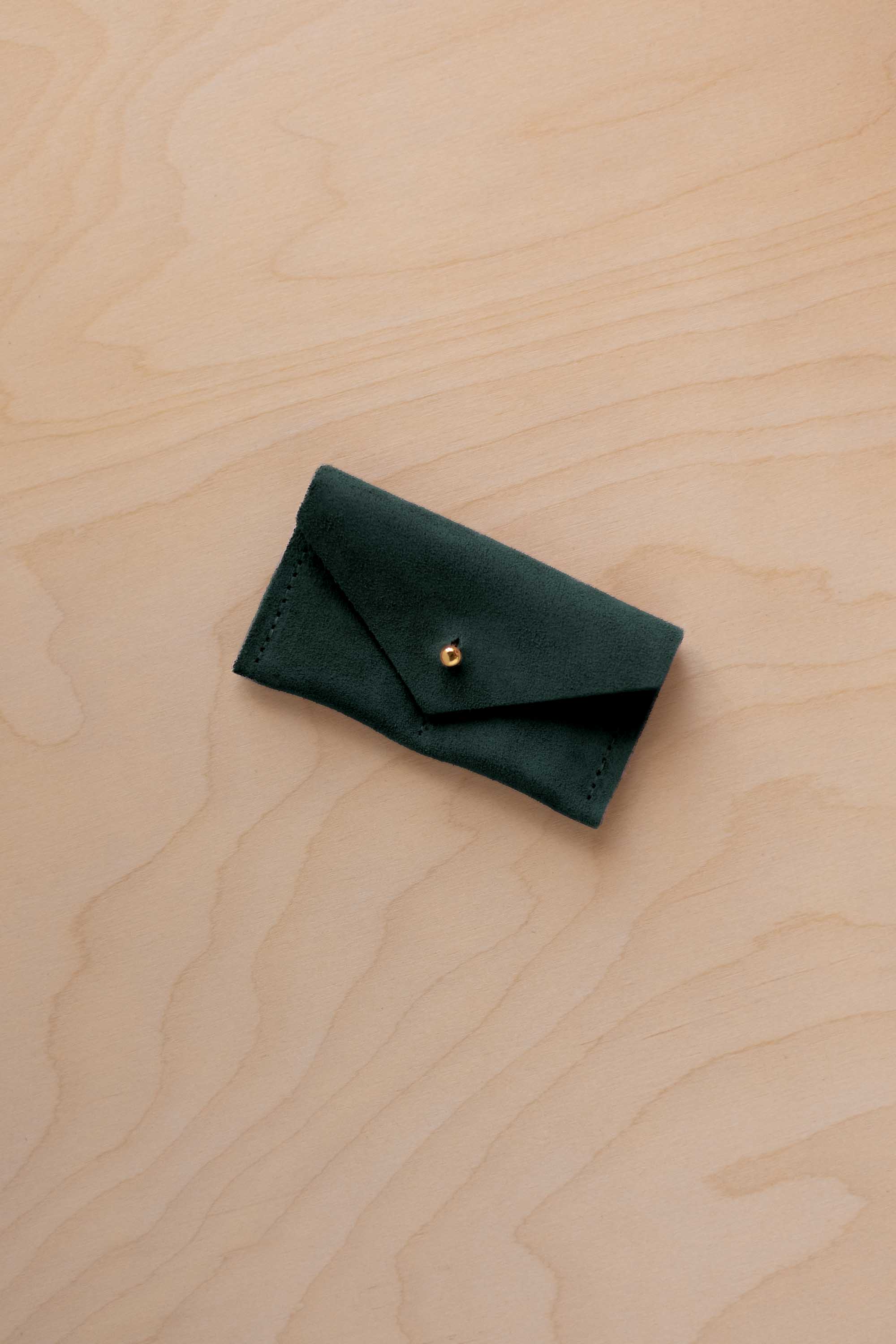 A small deep forest coloured suede jewellery pouch with a gold button, resting on a light wood grain surface.