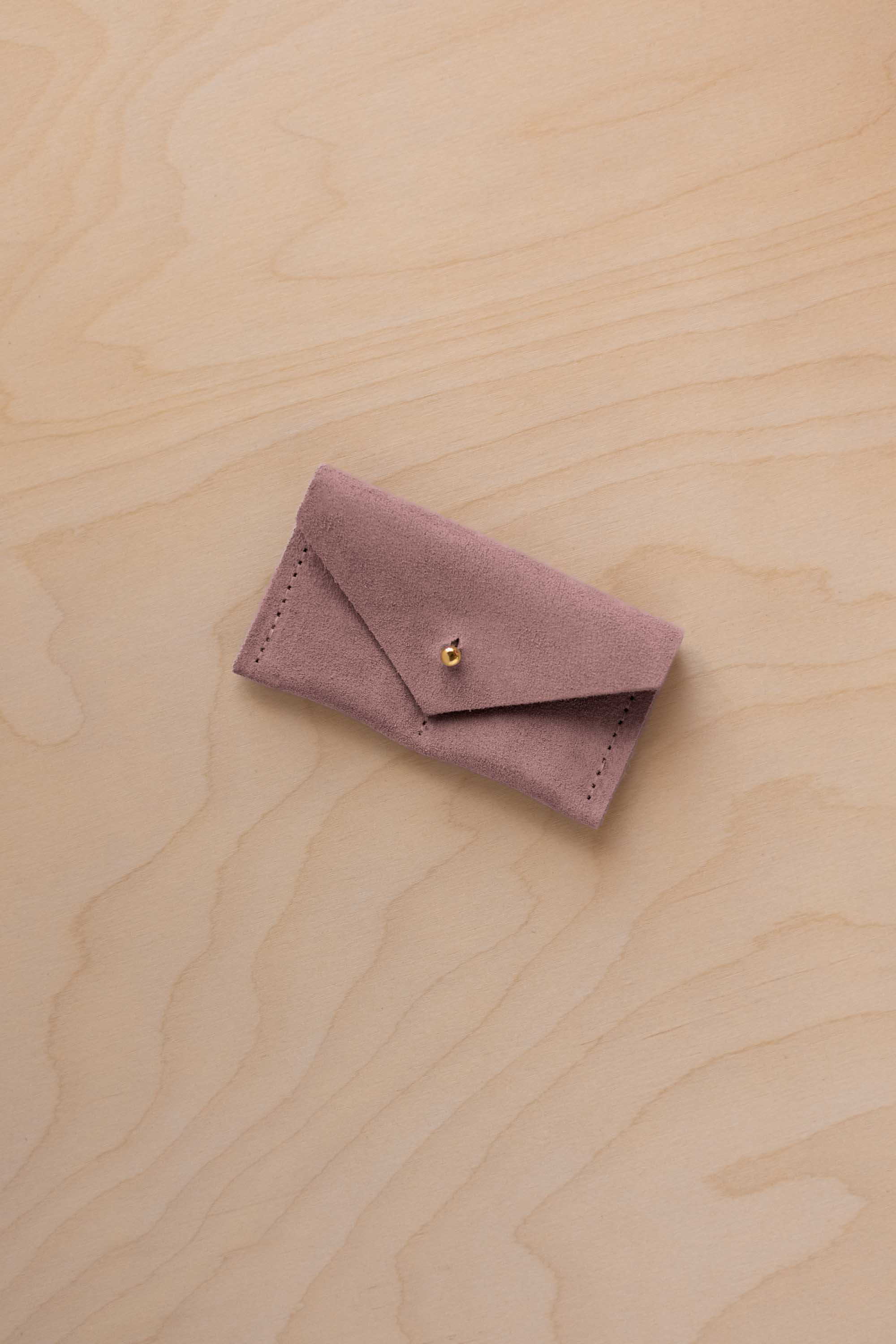 A small dusty mauve coloured suede jewellery pouch with a gold coloured fastening, resting on a light wood grain surface.