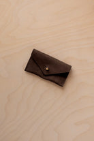 A small espresso brown coloured suede jewellery pouch with a gold button, resting on a light wood grain surface.