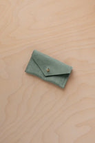 A small eucalyptus green coloured suede jewellery pouch with a gold coloured fastening, resting on a light wood grain surface.