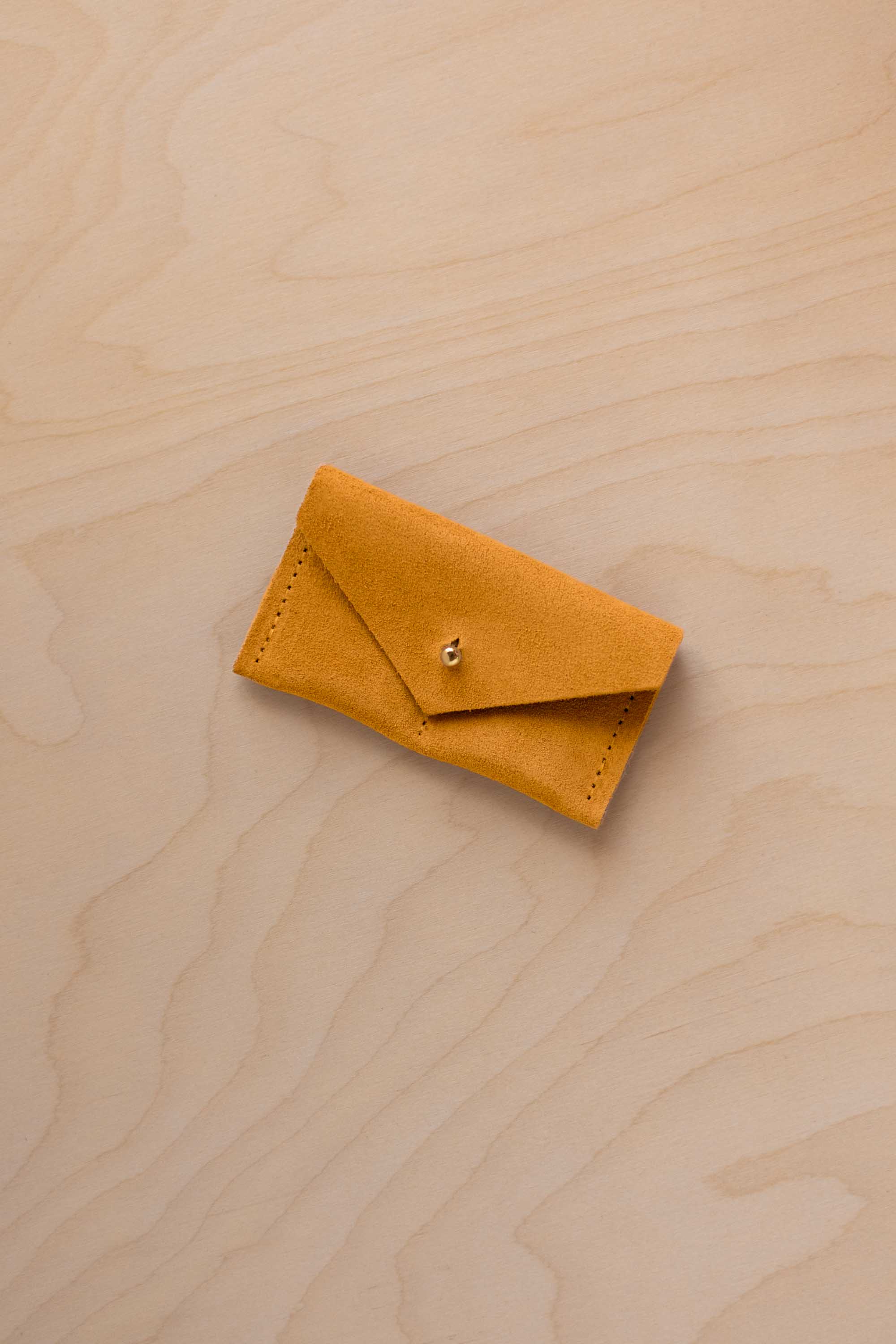 A small golden ochre coloured suede jewellery pouch with a gold coloured fastening, resting on a light wood grain surface.