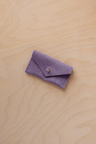A small lavender coloured suede jewellery pouch with a gold coloured fastening, resting on a light wood grain surface.