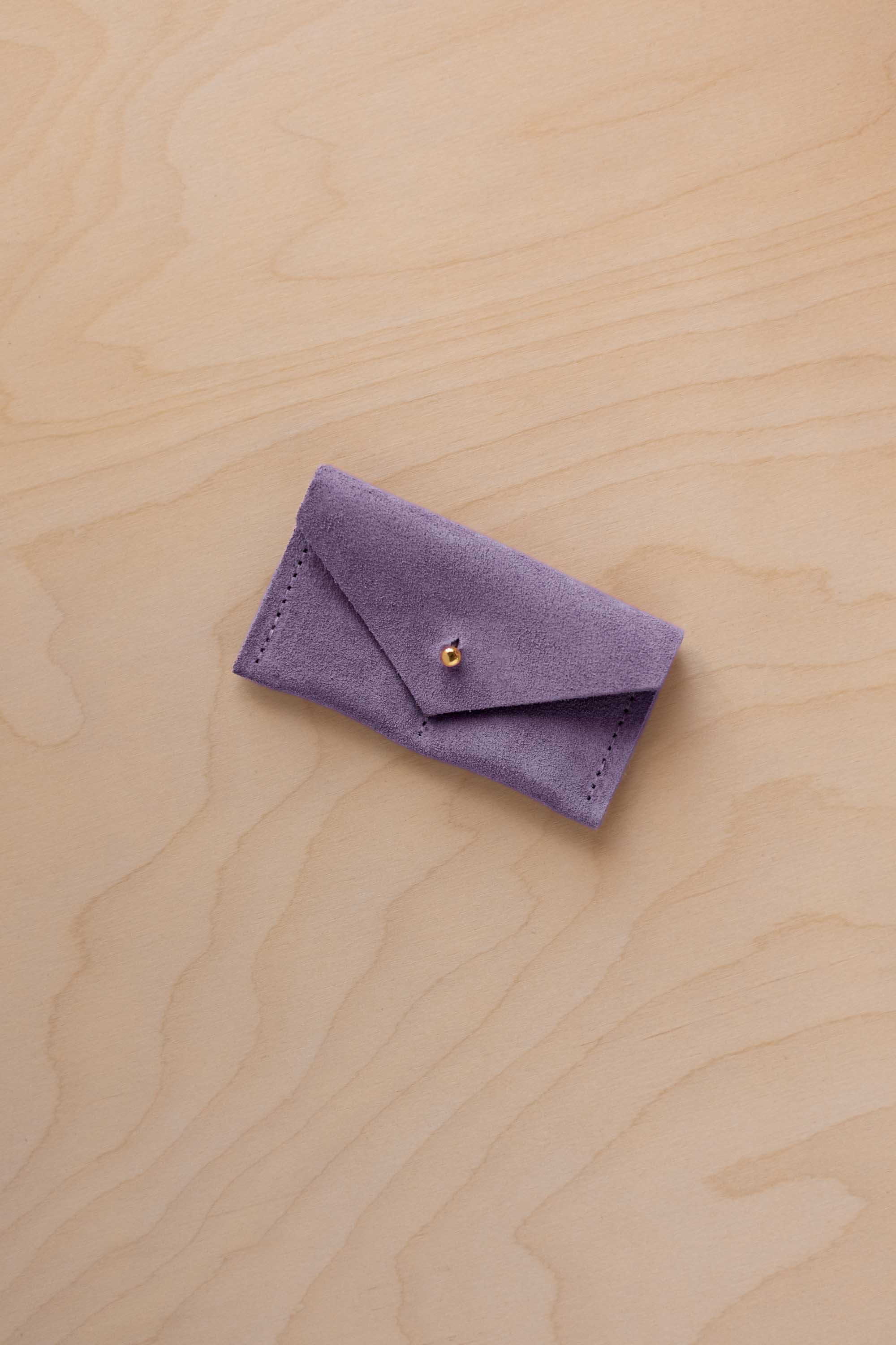 A small lavender coloured suede jewellery pouch with a gold coloured fastening, resting on a light wood grain surface.