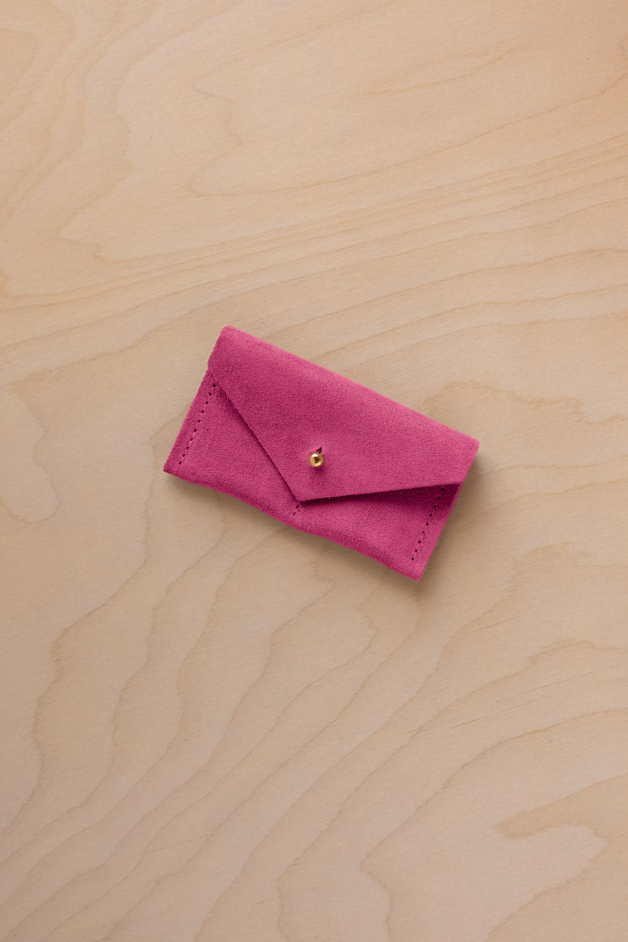 A small raspberry coloured suede jewellery pouch with a gold coloured fastening, resting on a light wood grain surface.