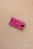 A small raspberry coloured suede jewellery pouch with a gold coloured fastening, resting on a light wood grain surface.