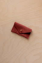 A small rust red coloured suede jewellery pouch with a gold coloured fastening, resting on a light wood grain surface.