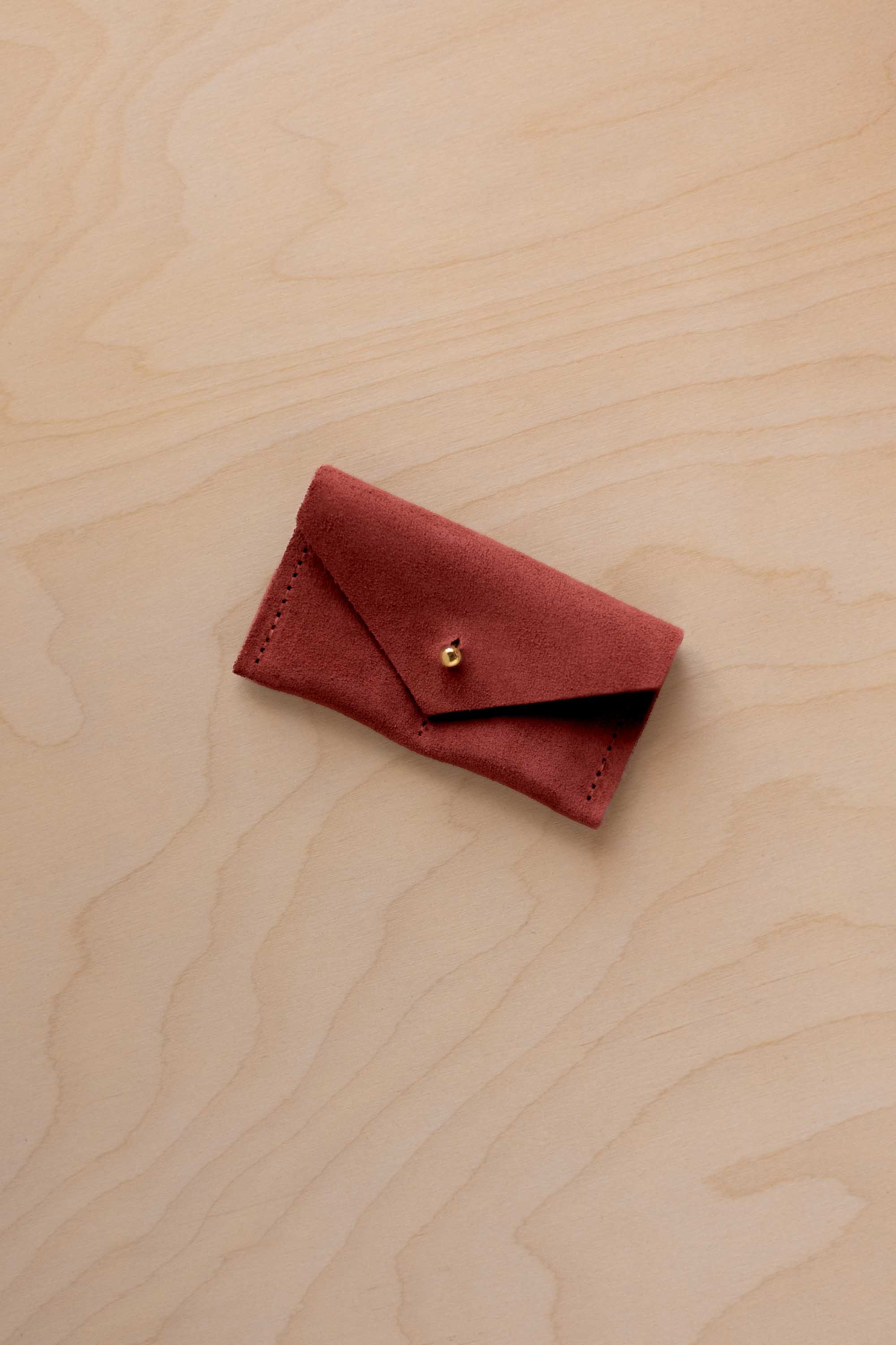 A small rust red coloured suede jewellery pouch with a gold coloured fastening, resting on a light wood grain surface.