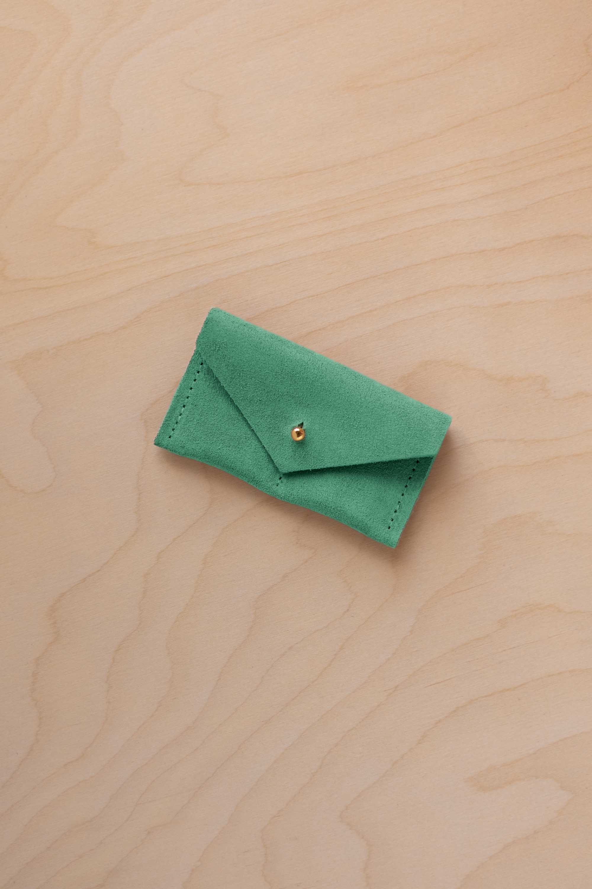 A small seafoam green coloured suede jewellery pouch with a gold coloured fastening, resting on a light wood grain surface.
