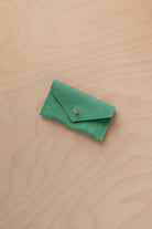 A small seafoam green coloured suede jewellery pouch with a gold coloured fastening, resting on a light wood grain surface.