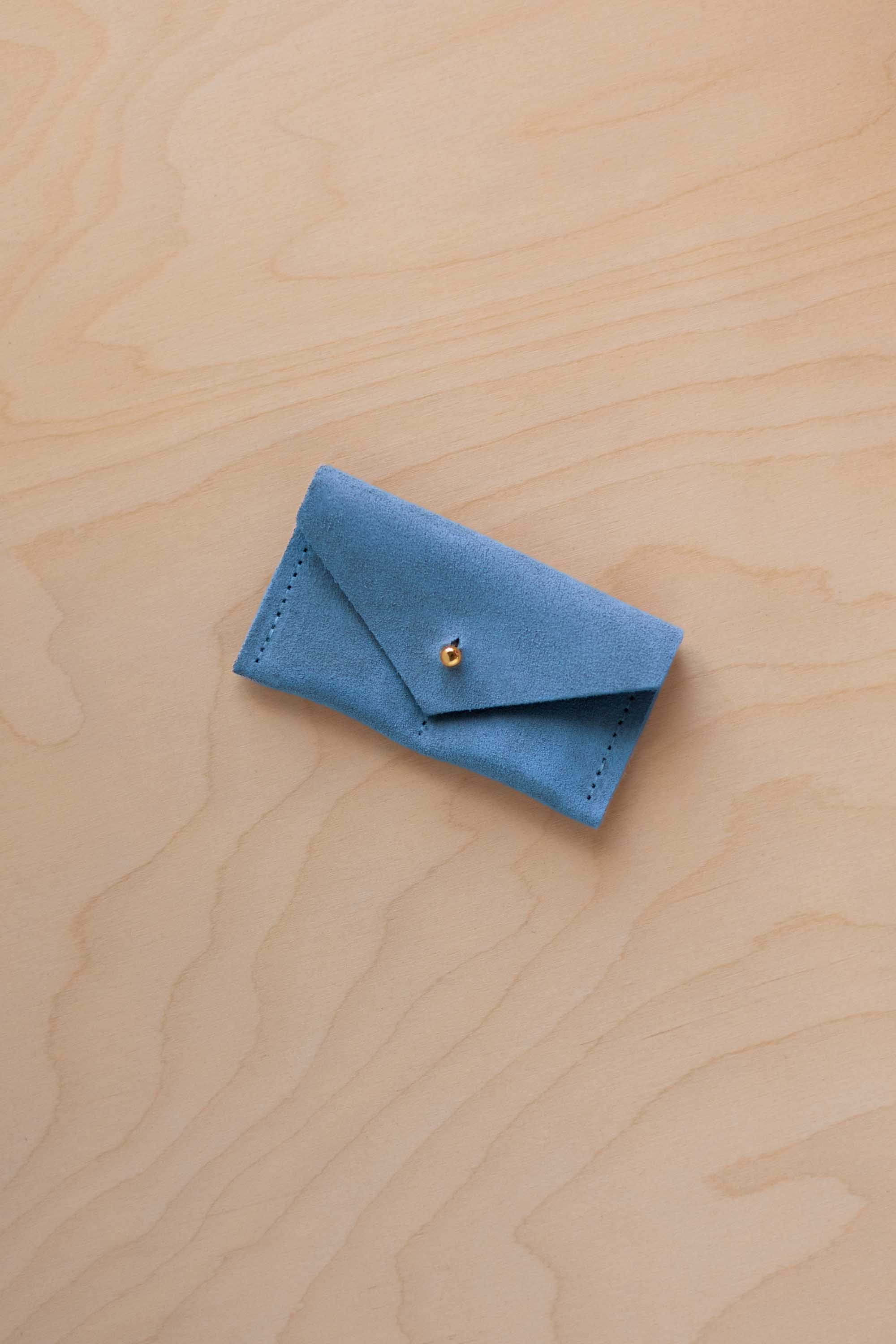 A small sky blue coloured suede jewellery pouch with a gold coloured fastening, resting on a light wood grain surface.
