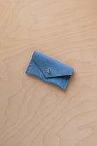 A small sky blue coloured suede jewellery pouch with a gold coloured fastening, resting on a light wood grain surface.