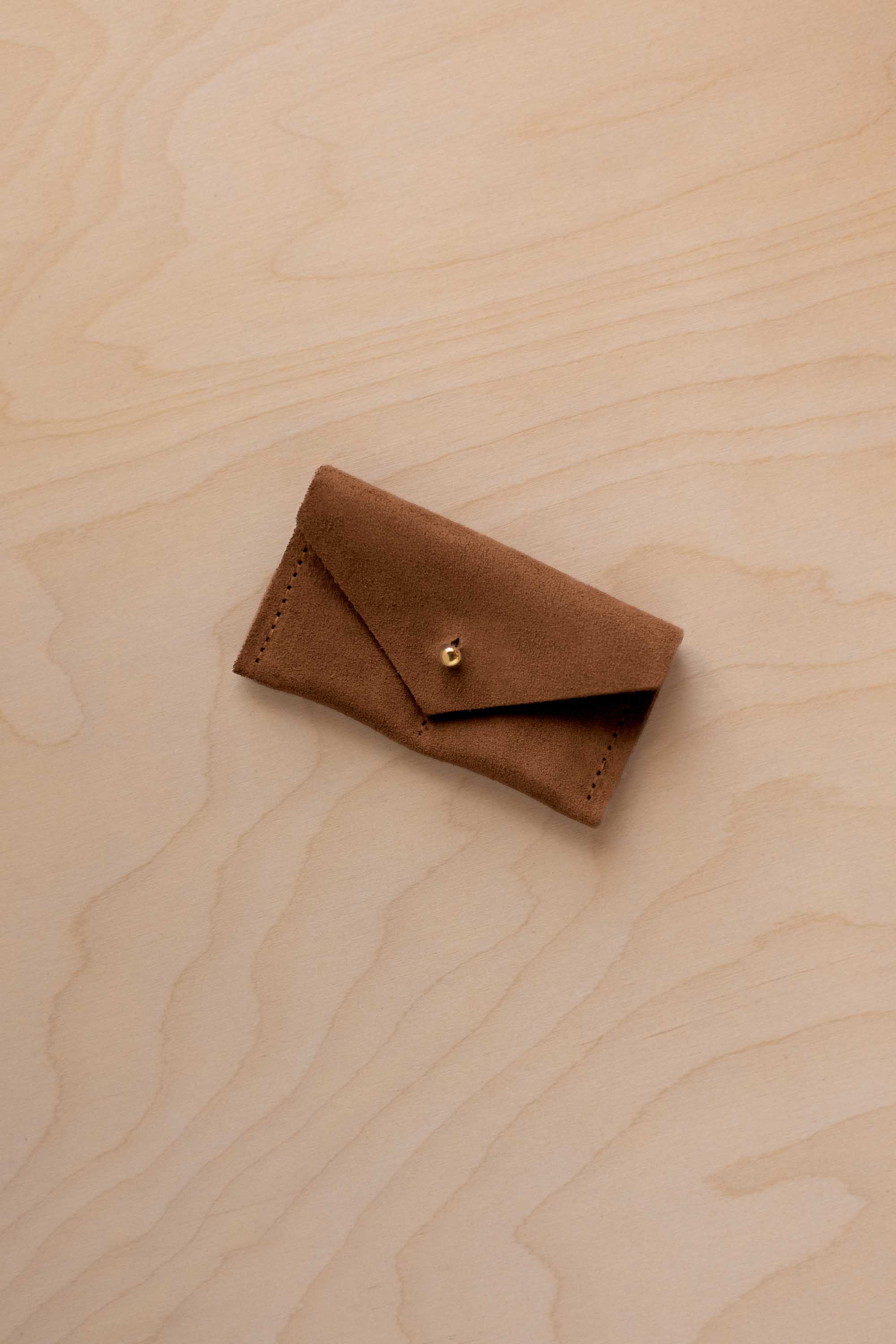 A small mocha brown coloured suede jewellery pouch with a gold coloured fastening, resting on a light wood grain surface.