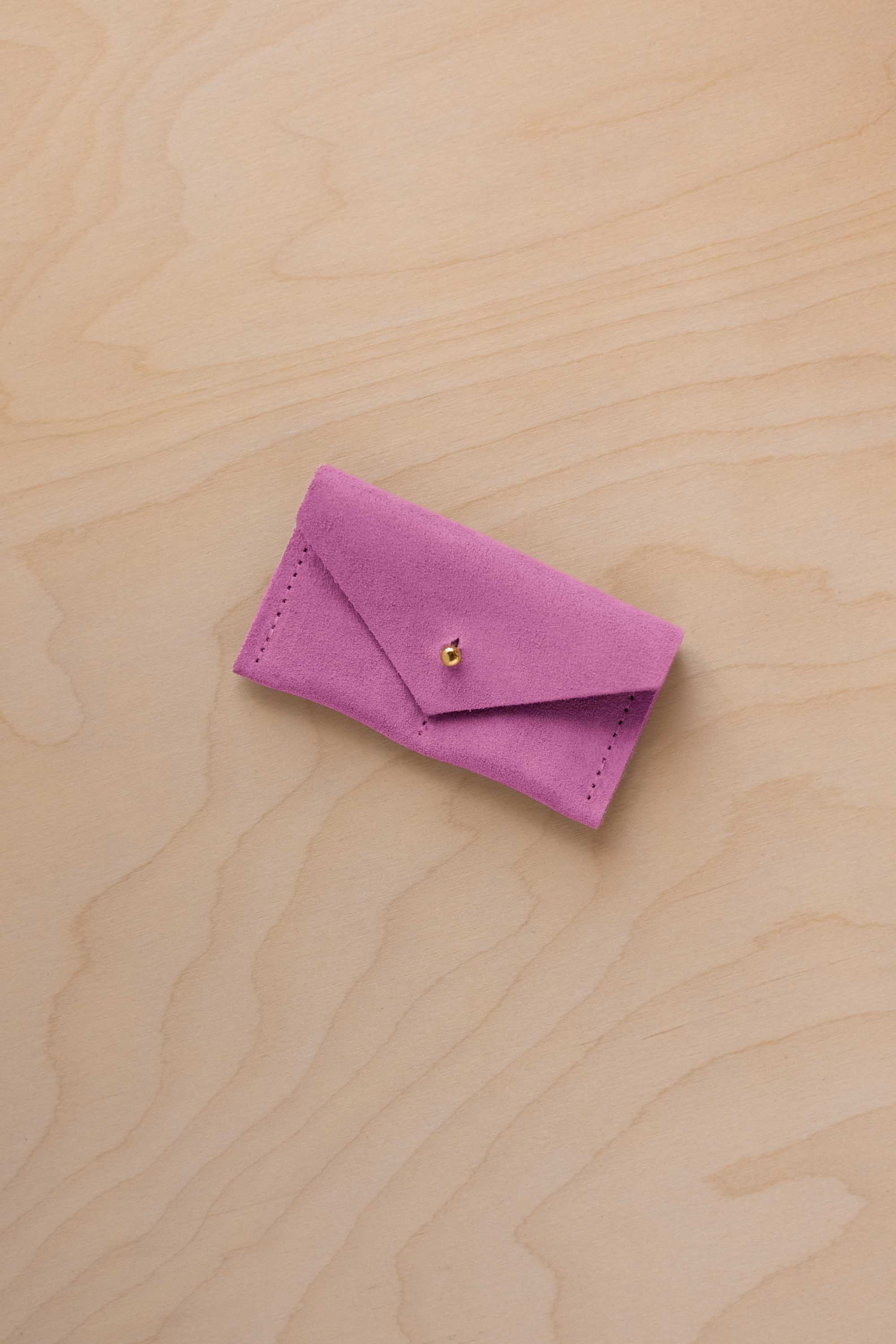 A small peony pink coloured suede jewellery pouch with a gold coloured fastening, resting on a light wood grain surface.