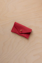 A small scarlet red coloured suede jewellery pouch with a gold coloured fastening, resting on a light wood grain surface.