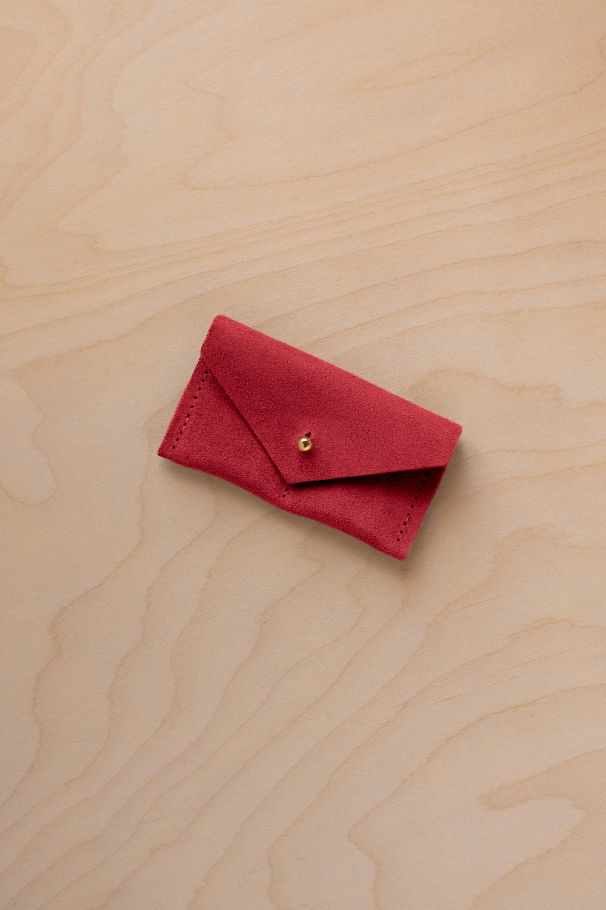 A small scarlet red coloured suede jewellery pouch with a gold coloured fastening, resting on a light wood grain surface.