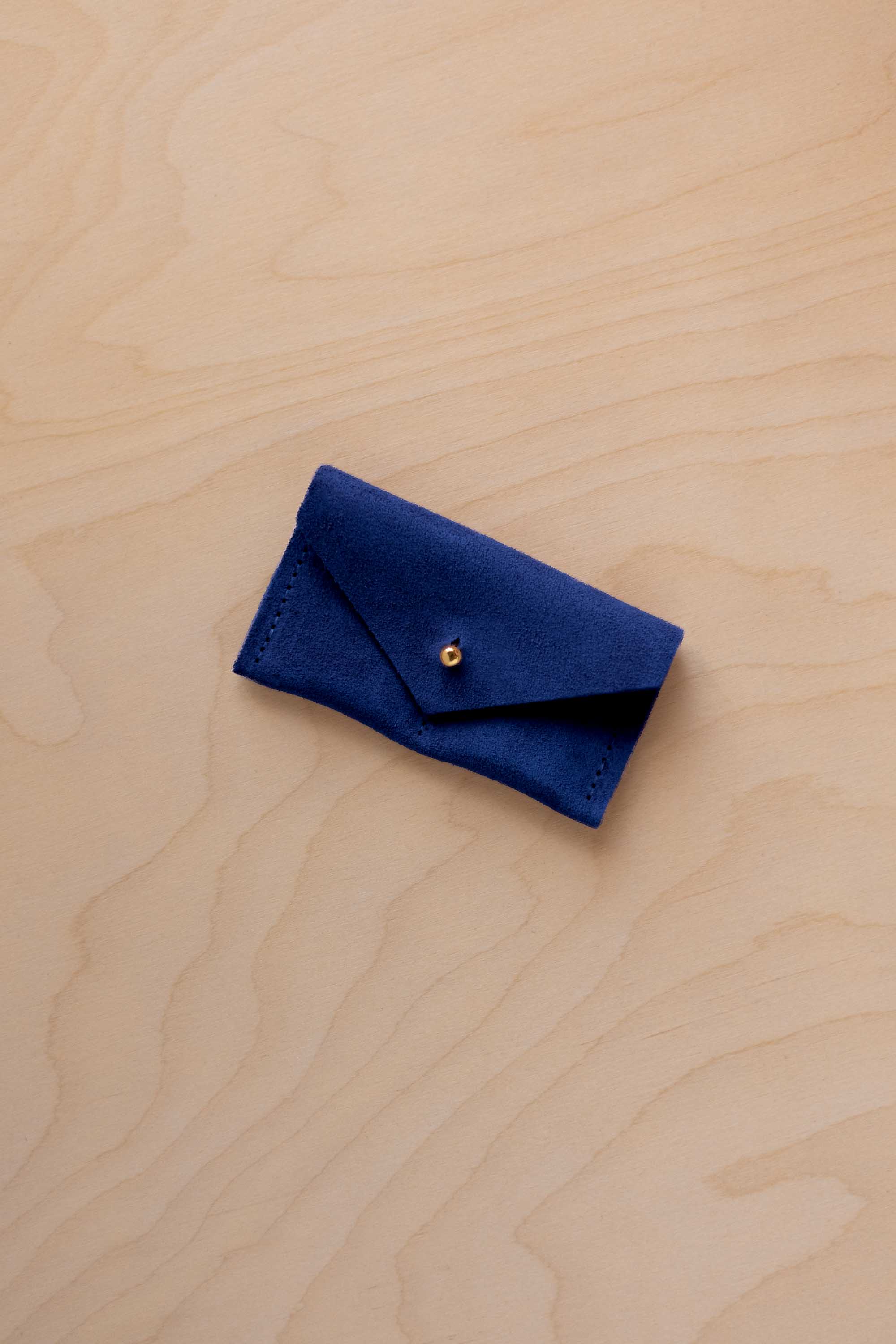 A small ultramarine blue coloured suede jewellery pouch with a gold coloured fastening, resting on a light wood grain surface.