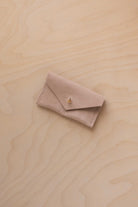 A small warm sand coloured suede jewellery pouch with a gold coloured fastening, resting on a light wood grain surface.