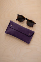 A pair of dark brown tortoiseshell sunglasses with round frames, resting on a light beige wood-grain surface next to a blackcurrant dark purple coloured suede glasses case with gold coloured buttons.